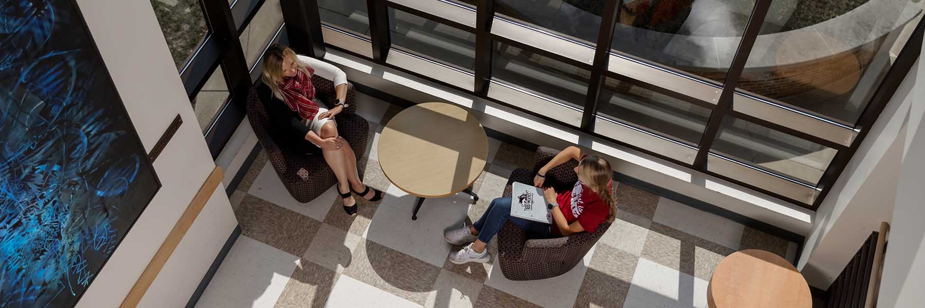 A faculty member meeting with a student next to large windows